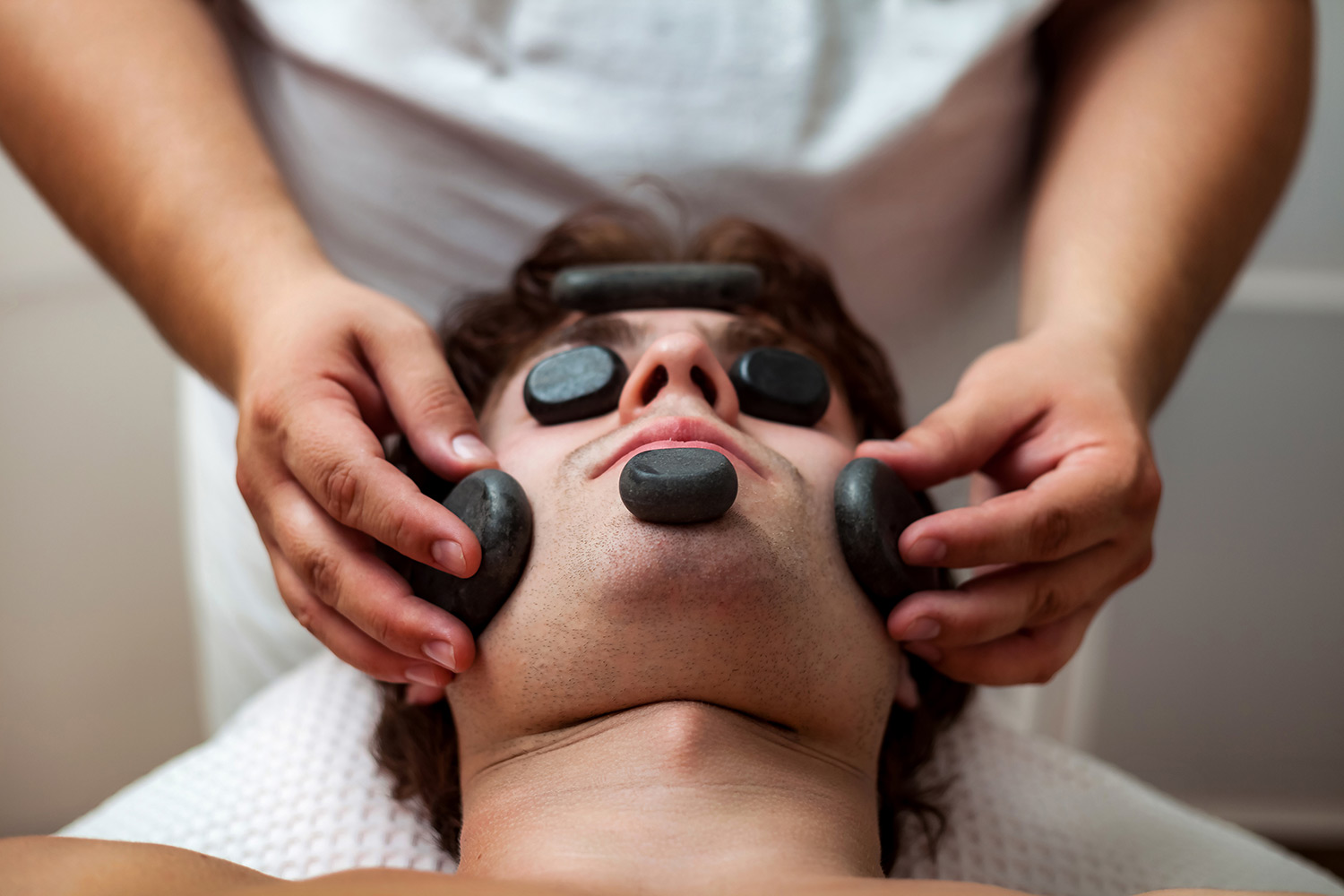 Man receiving a hot stone facial treatment with smooth stones placed on his face, surrounded by a therapist's hands, promoting relaxation and wellness through the Thermal Stone Stretch Ritual.