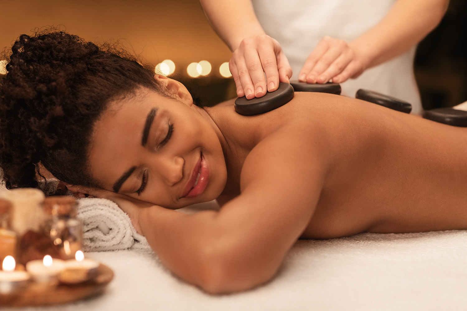 Female receiving hot stone massage with black stones on her back, surrounded by spa elements and soft candlelight, illustrating relaxation and wellness benefits.